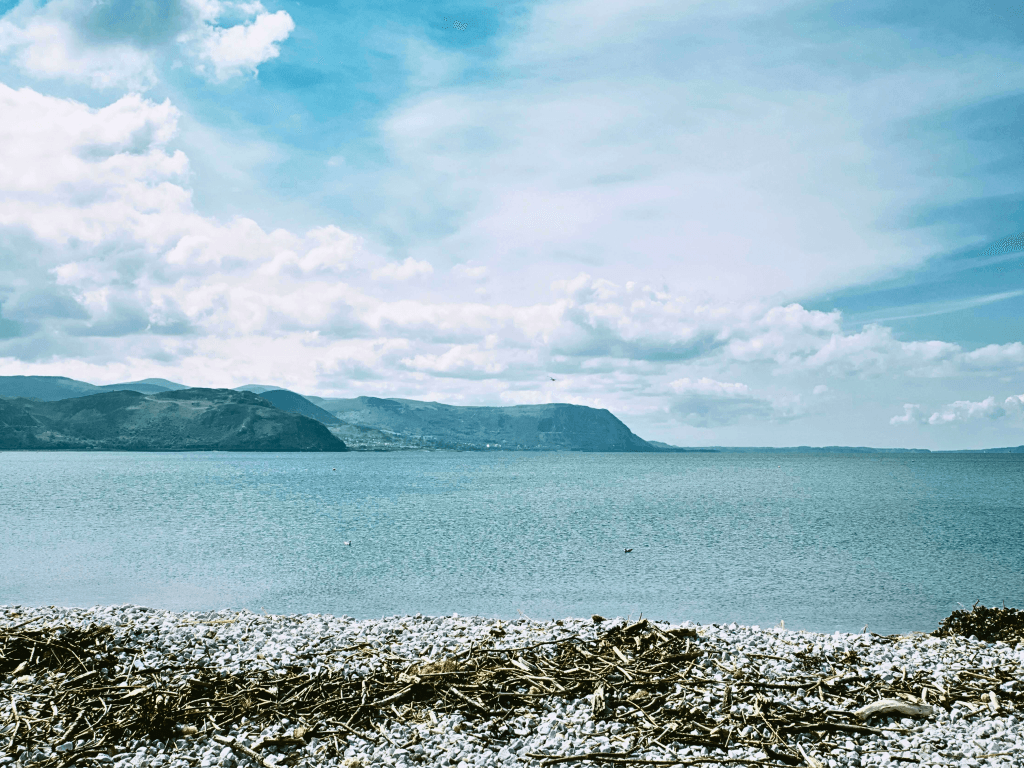 Peaceful Welsh beach with pebbled shore, calm blue sea, and distant hills under a partly cloudy sky - an ideal spot for quiet hidden beaches for family escapes