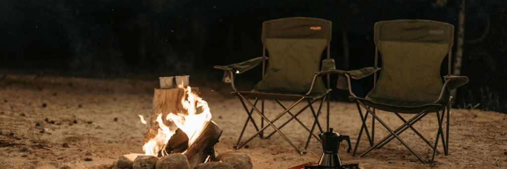 Two empty camping chairs beside a glowing campfire on a quiet UK campsite at night. 