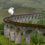Steam train crossing viaduct in the Scottish Highlands, surrounded by lush greenery and misty hills.